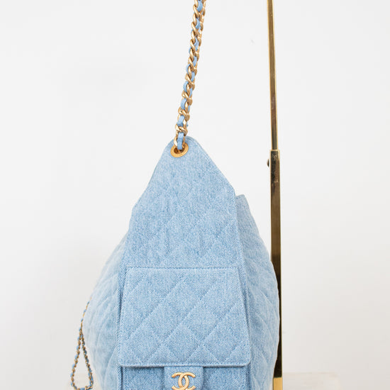 A light blue quilted denim shoulder bag with gold-tone hardware, displayed on a white surface next to a gold metal stand against a white background.