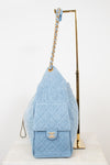 A light blue quilted denim shoulder bag with gold-tone hardware, displayed on a white surface next to a gold metal stand against a white background.