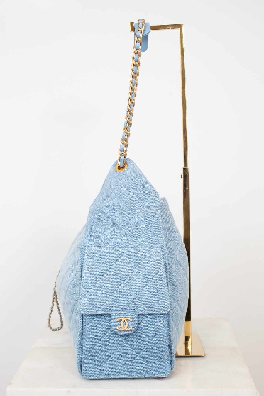 A light blue quilted denim shoulder bag with gold-tone hardware, displayed on a white surface next to a gold metal stand against a white background.