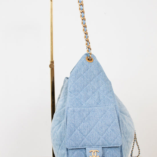 A light blue quilted Chanel shoulder bag made of textured canvas, displayed on a gold metal stand against a white background.