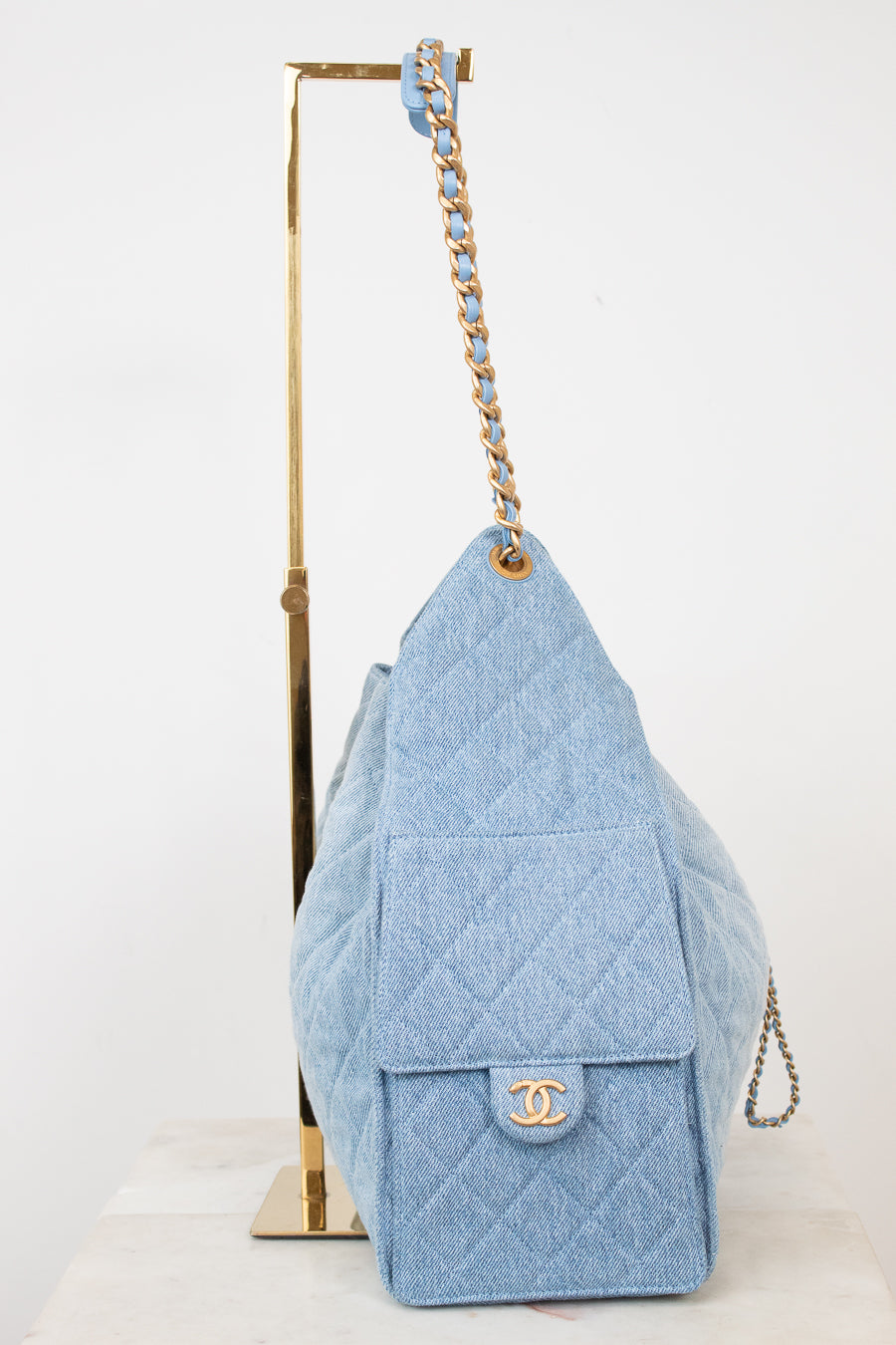 A light blue quilted Chanel shoulder bag made of textured canvas, displayed on a gold metal stand against a white background.