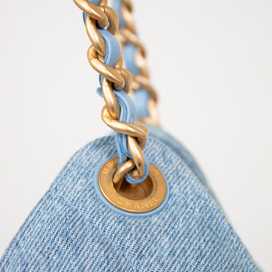 Close-up of a light blue denim shoulder bag with a gold-tone chain strap and a matching gold-tone grommet, set against a clean white background.