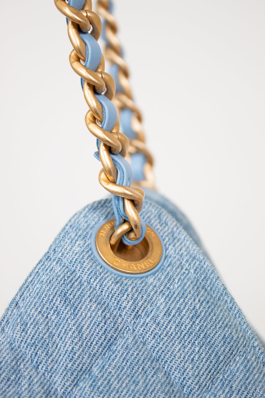 Close-up of a light blue denim shoulder bag with a gold-tone chain strap and a matching gold-tone grommet, set against a clean white background.