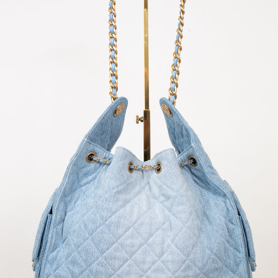 A light blue quilted denim drawstring handbag with gold-tone chain shoulder straps, displayed on a white background with a gold stand.
