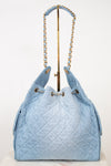 A light blue quilted denim drawstring handbag with gold-tone chain shoulder straps, displayed on a white background with a gold stand.