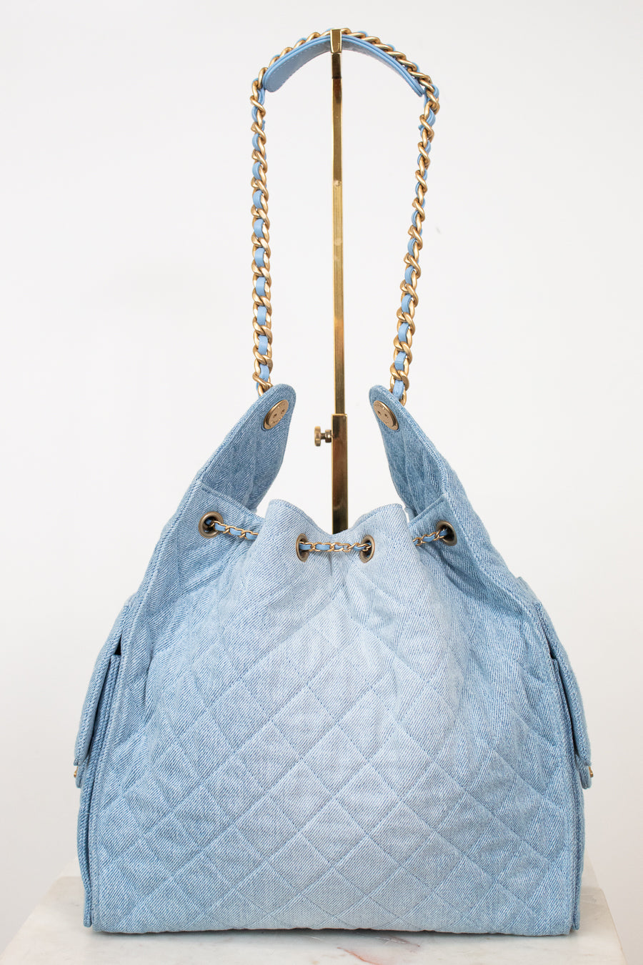 A light blue quilted denim drawstring handbag with gold-tone chain shoulder straps, displayed on a white background with a gold stand.