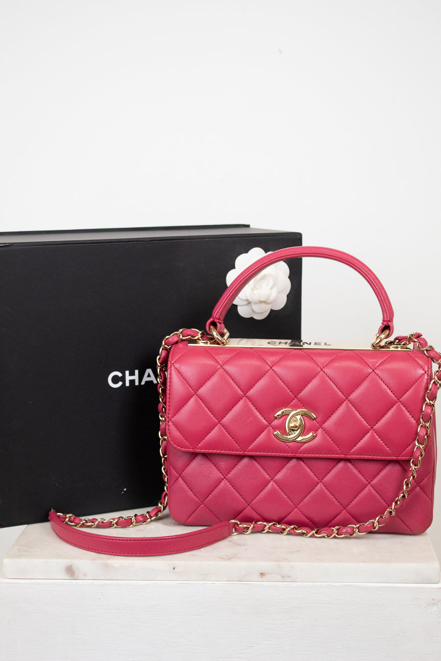 Pink quilted handbag with gold chain and clasp on a white surface with a black box in the background.