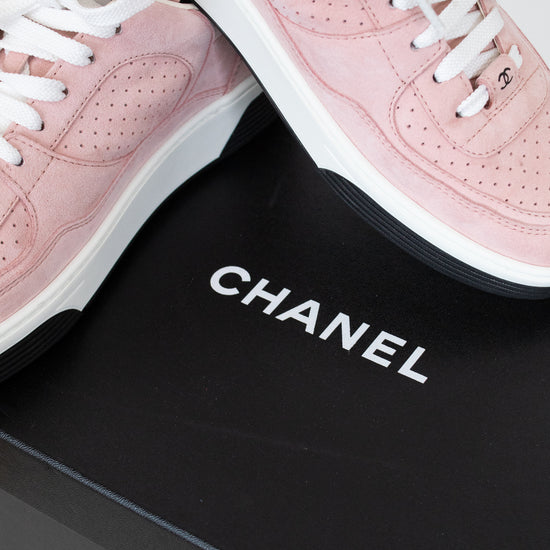 A pair of Chanel low-top sneakers in light pink suede with white laces and white midsoles, displayed on a black Chanel shoebox.