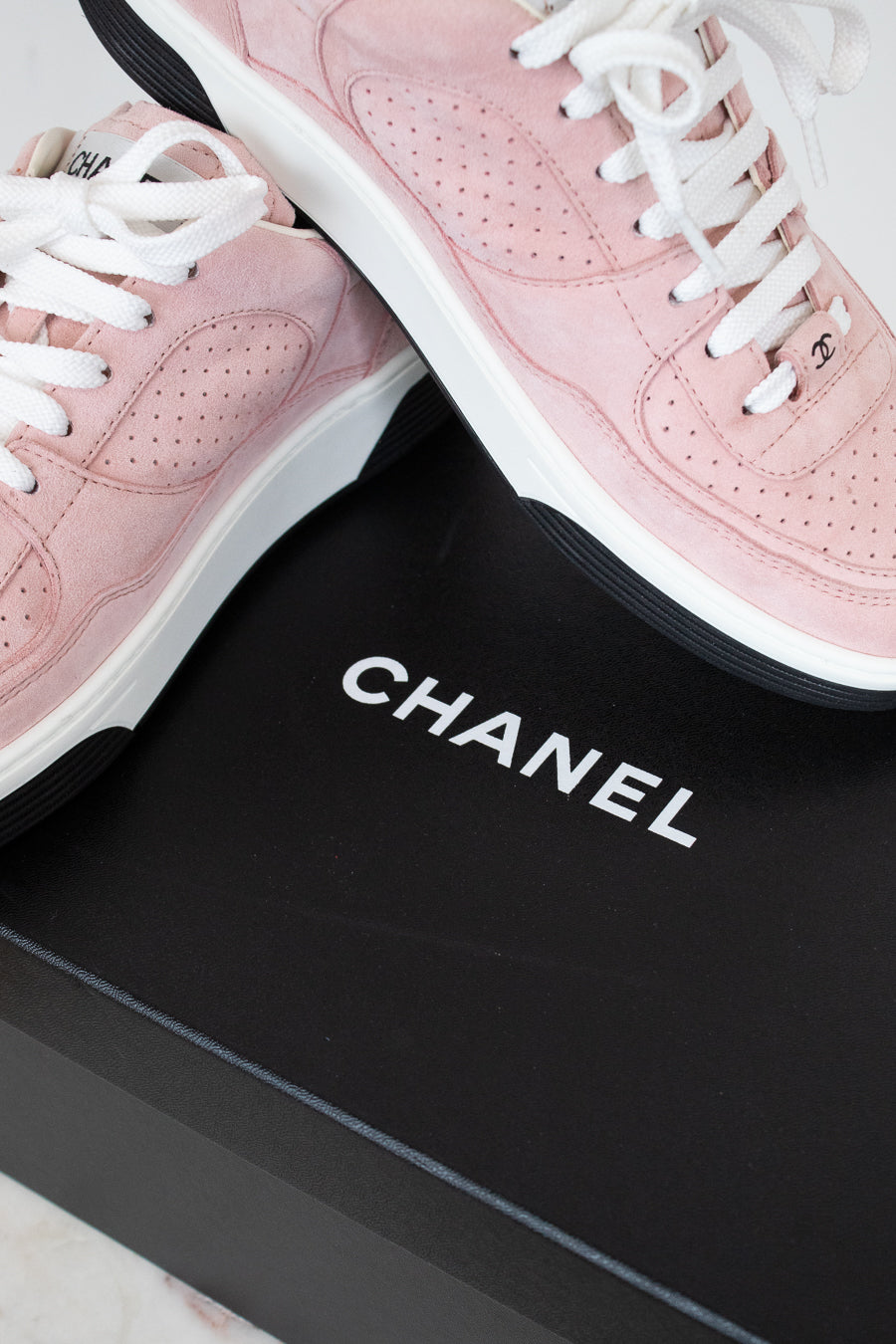 A pair of Chanel low-top sneakers in light pink suede with white laces and white midsoles, displayed on a black Chanel shoebox.