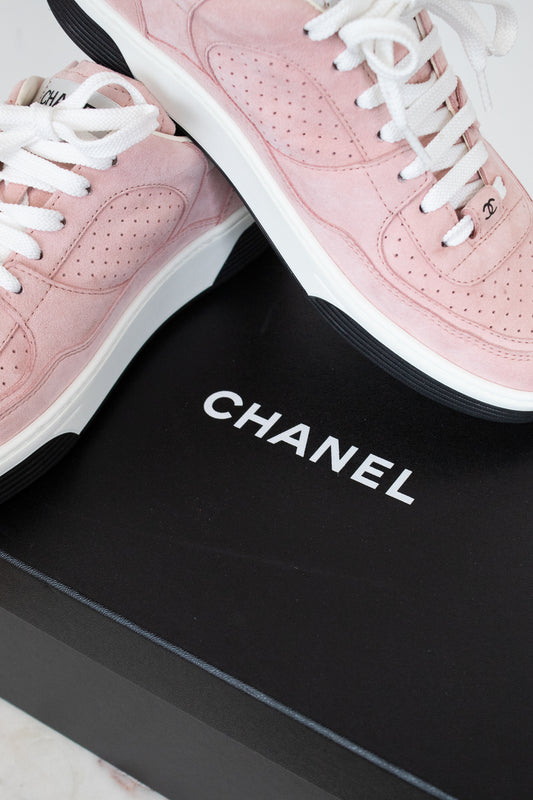 A pair of Chanel low-top sneakers in light pink suede with white laces and white midsoles, displayed on a black Chanel shoebox.