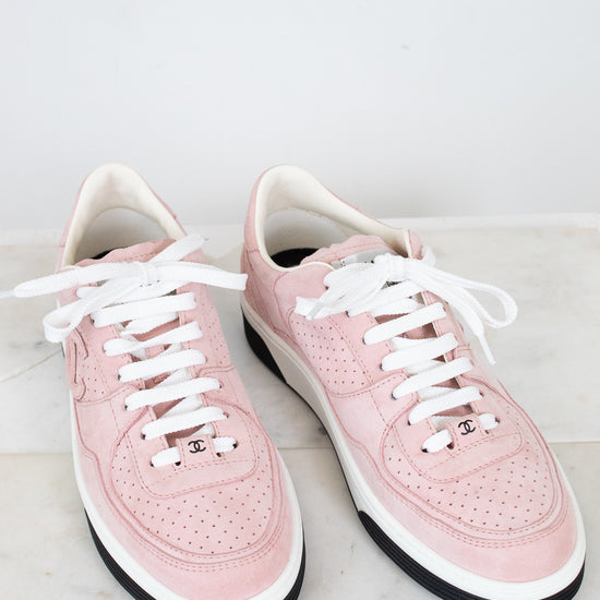 A pair of light pink leather low-top sneakers with white laces, white midsoles, and black outsoles, displayed on a light marble surface against a white background.