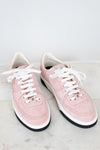 A pair of light pink leather low-top sneakers with white laces, white midsoles, and black outsoles, displayed on a light marble surface against a white background.