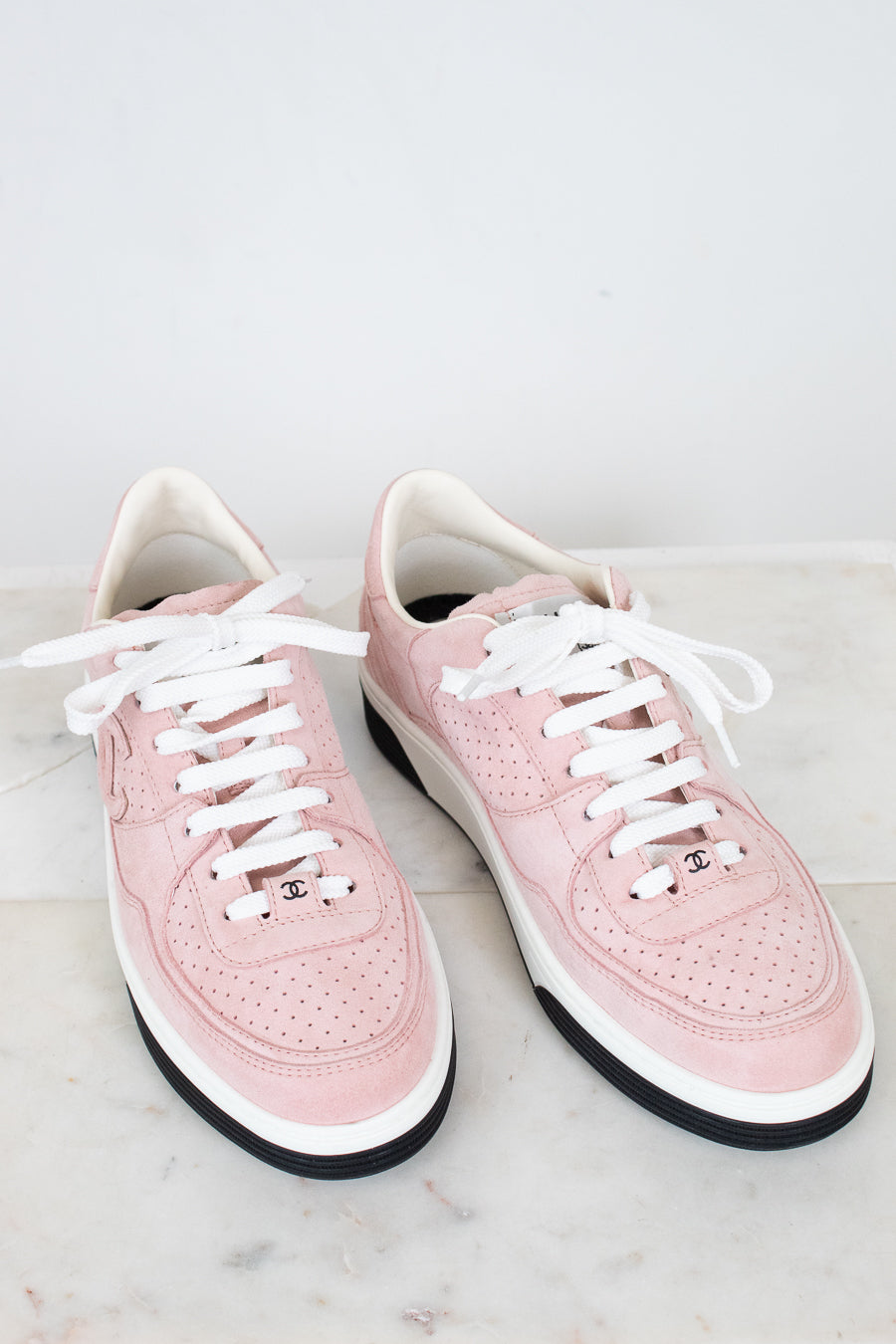 A pair of light pink leather low-top sneakers with white laces, white midsoles, and black outsoles, displayed on a light marble surface against a white background.
