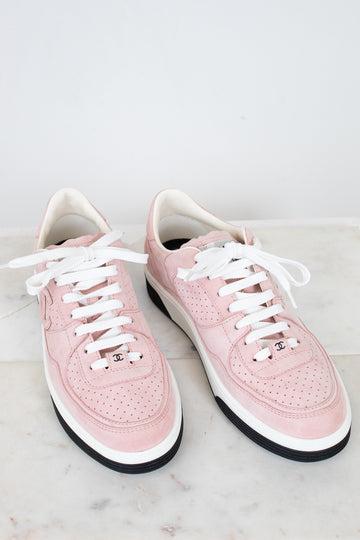 A pair of light pink leather low-top sneakers with white laces, white midsoles, and black outsoles, displayed on a light marble surface against a white background.