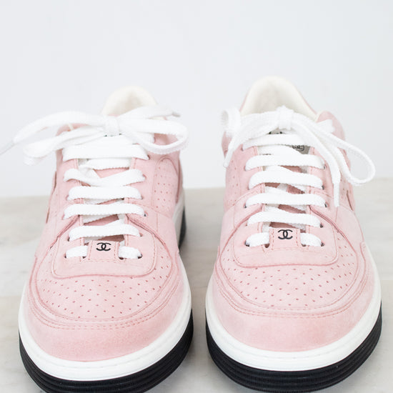 A pair of low-top Chanel sneakers in light pink suede and leather, featuring white laces, white midsoles, black outsoles, and the iconic interlocking CC logo on the tongue.