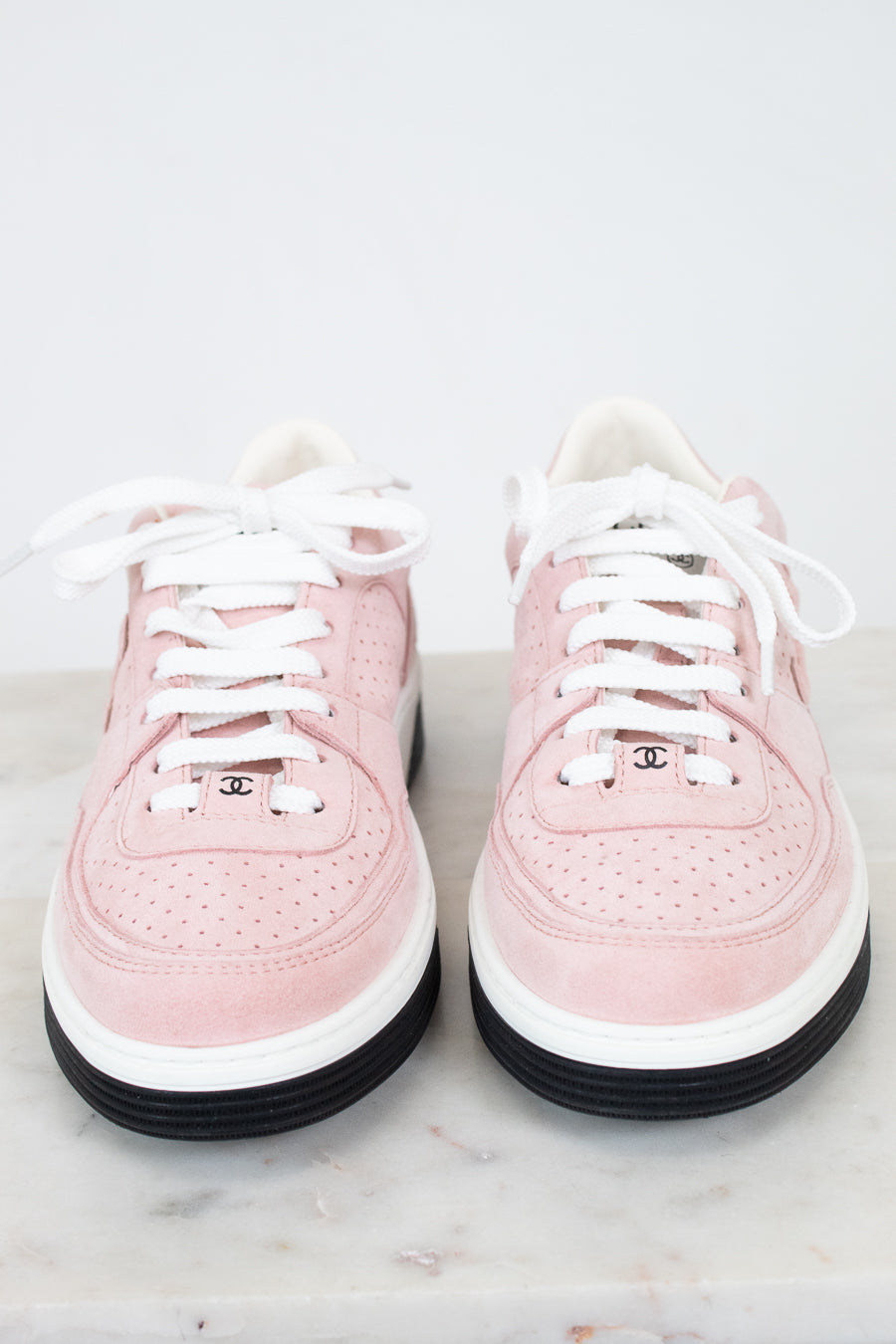A pair of low-top Chanel sneakers in light pink suede and leather, featuring white laces, white midsoles, black outsoles, and the iconic interlocking CC logo on the tongue.
