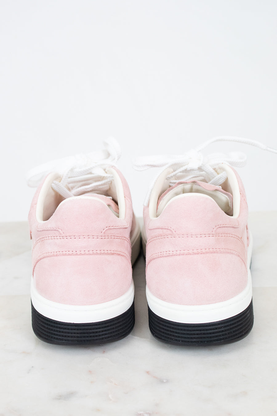 A pair of low-top sneakers shown from the back, featuring light pink suede uppers, white laces, a white midsole, and a black rubber outsole, set against a clean white background.