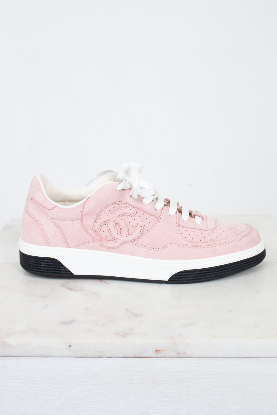 A light pink Chanel low-top sneaker with white laces, a white midsole, and a black outsole, displayed on a white marble surface against a white background.