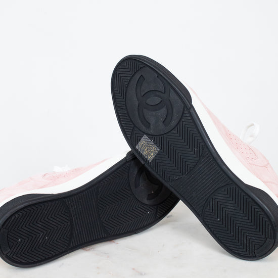 A pair of Chanel low-top sneakers shown from the bottom, highlighting the black textured rubber soles with the embossed interlocking CC logo. The uppers are light pink with white side panels, resting on a white marble surface against a white background.