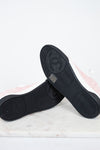 A pair of Chanel low-top sneakers shown from the bottom, highlighting the black textured rubber soles with the embossed interlocking CC logo. The uppers are light pink with white side panels, resting on a white marble surface against a white background.