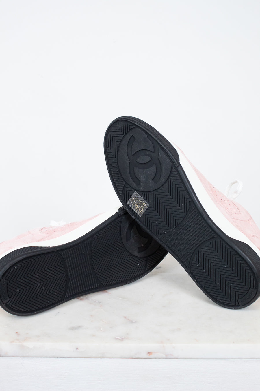 A pair of Chanel low-top sneakers shown from the bottom, highlighting the black textured rubber soles with the embossed interlocking CC logo. The uppers are light pink with white side panels, resting on a white marble surface against a white background.