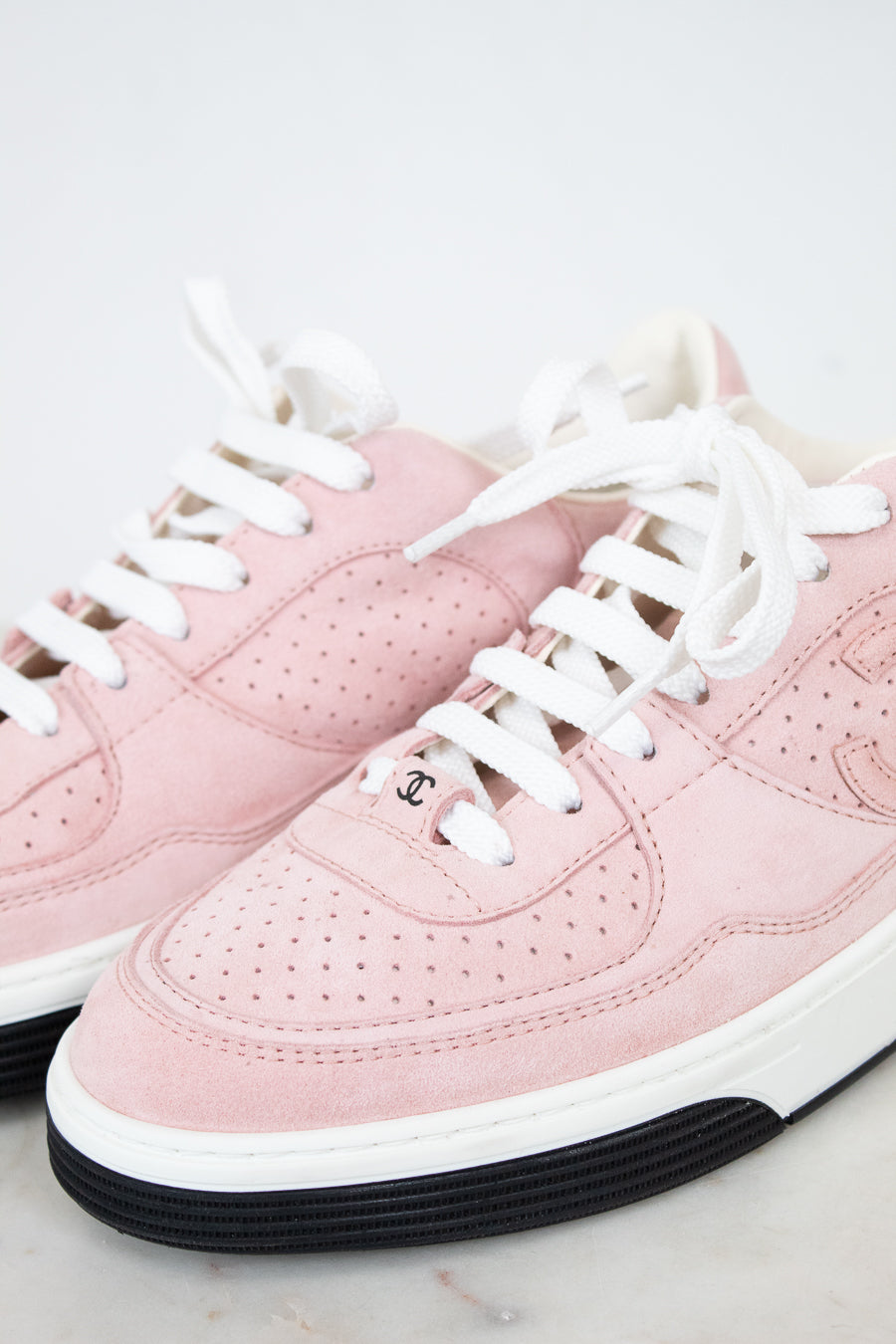 A pair of low-top pink sneakers with white laces, a white midsole, and a black rubber outsole. One shoe shows a small interlocking CC logo on the side, indicating they are Chanel brand.