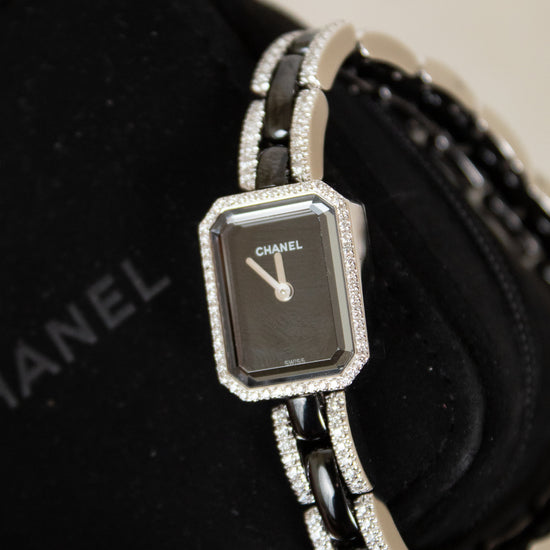 Chanel watch with a silver-toned rectangular face, black dial, and a silver-and-black link bracelet embellished with diamonds, displayed on a black Chanel box.