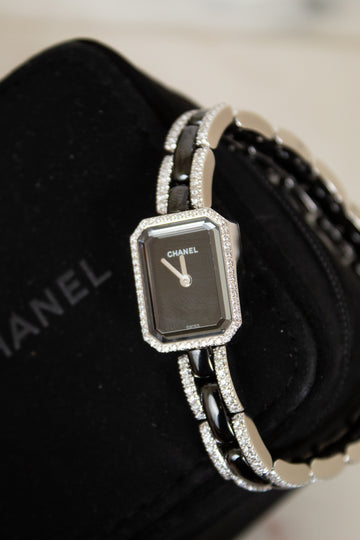 Chanel watch with a silver-toned rectangular face, black dial, and a silver-and-black link bracelet embellished with diamonds, displayed on a black Chanel box.
