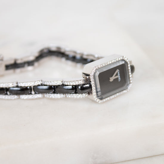 A close-up of a Chanel watch with a black dial, silver-tone link details, and a bracelet featuring black links and pavé-set diamonds, resting on a white marble surface.