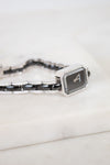 A close-up of a Chanel watch with a black dial, silver-tone link details, and a bracelet featuring black links and pavé-set diamonds, resting on a white marble surface.