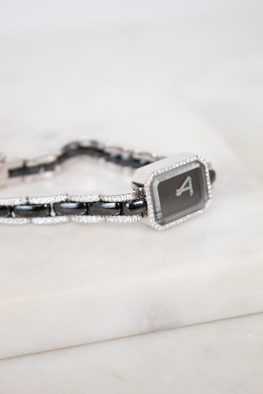 A close-up of a Chanel watch with a black dial, silver-tone link details, and a bracelet featuring black links and pavé-set diamonds, resting on a white marble surface.