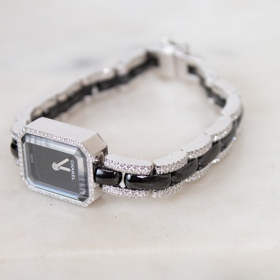 A women's watch with a rectangular silver-tone face, a black and silver link, and a bracelet featuring alternating black links and pavé-set diamonds, resting on a light marble surface.