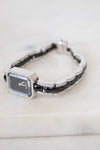 A women's watch with a rectangular silver-tone face, a black and silver link, and a bracelet featuring alternating black links and pavé-set diamonds, resting on a light marble surface.