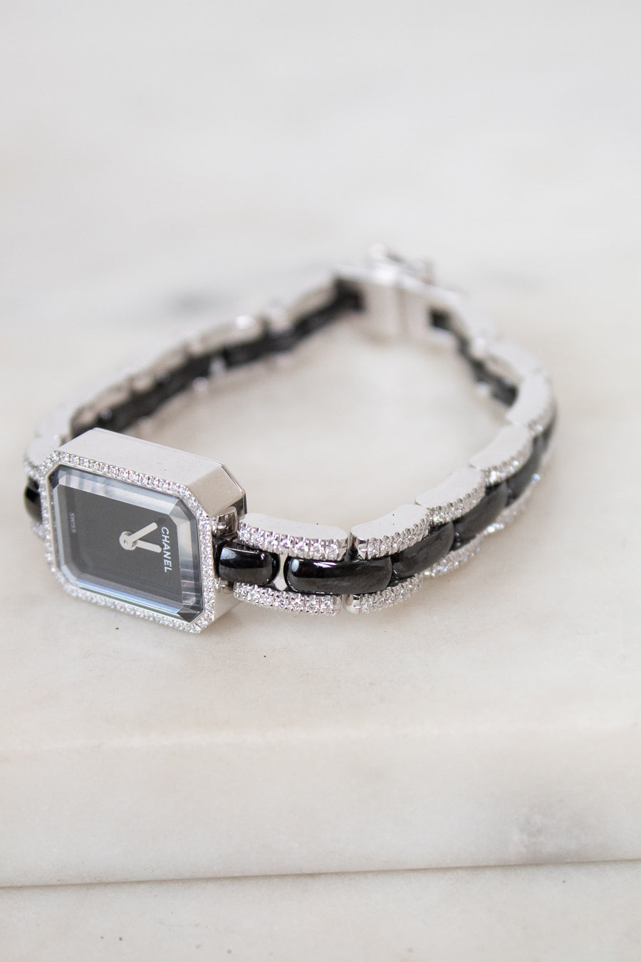 A women's watch with a rectangular silver-tone face, a black and silver link, and a bracelet featuring alternating black links and pavé-set diamonds, resting on a light marble surface.