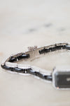 A close-up of a bracelet with alternating glossy black and polished silver links, accented with small diamonds, resting on a light marble surface.