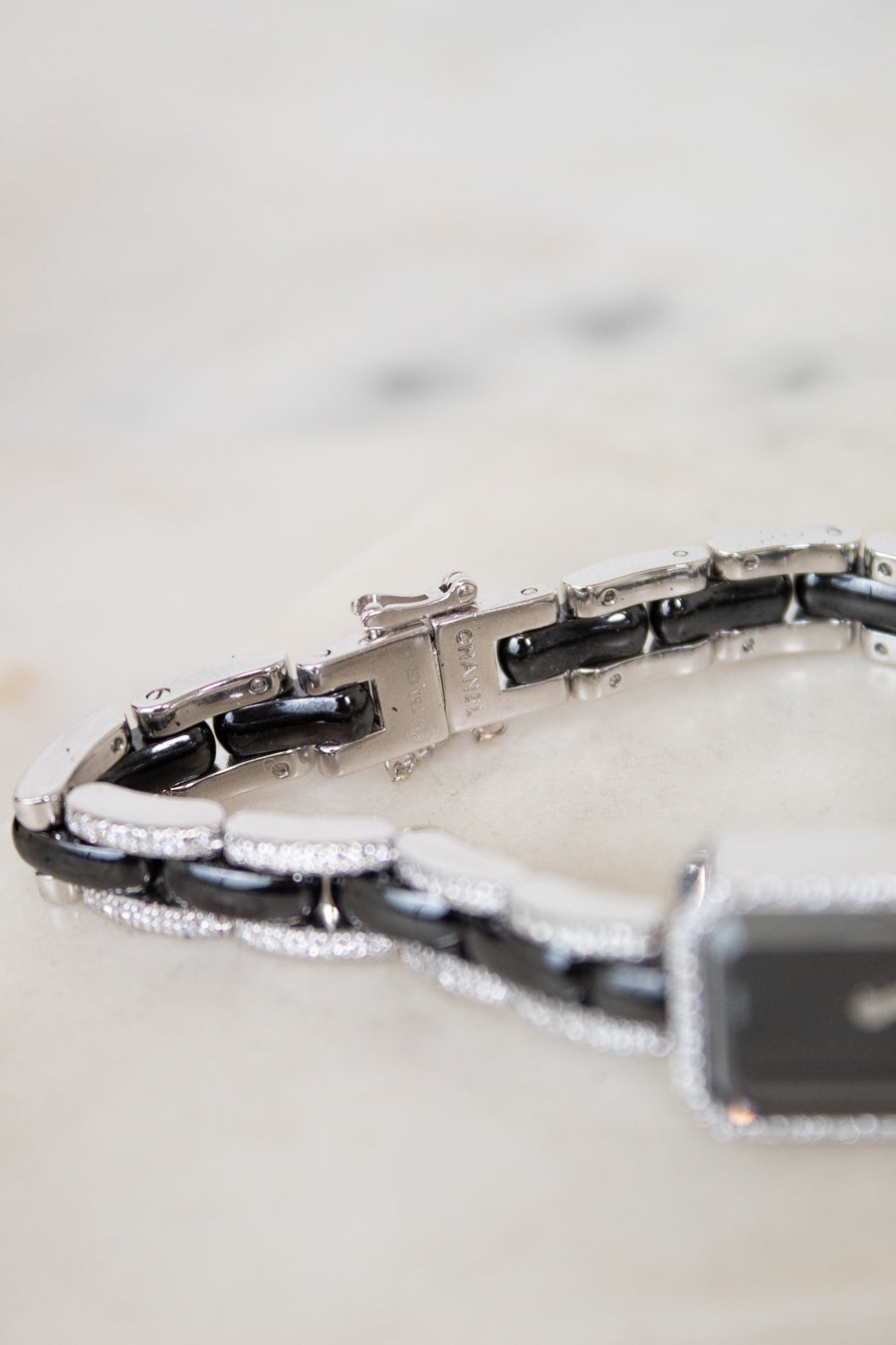 A close-up of a bracelet with alternating glossy black and polished silver links, accented with small diamonds, resting on a light marble surface.