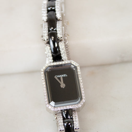 A Chanel watch with a black rectangular face and a silver-tone bracelet paved with clear stones, resting on a light marble surface.