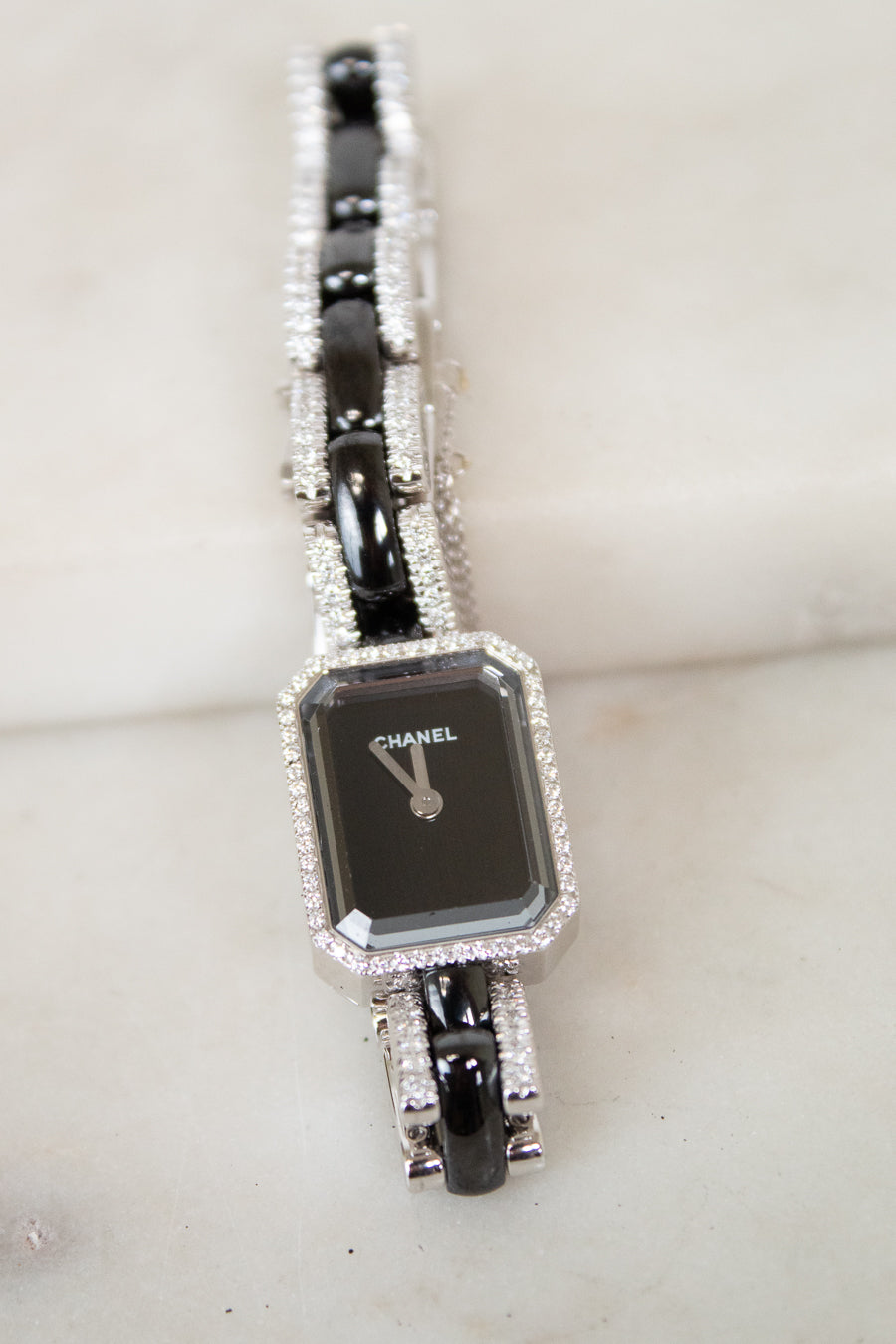 A Chanel watch with a black rectangular face and a silver-tone bracelet paved with clear stones, resting on a light marble surface.