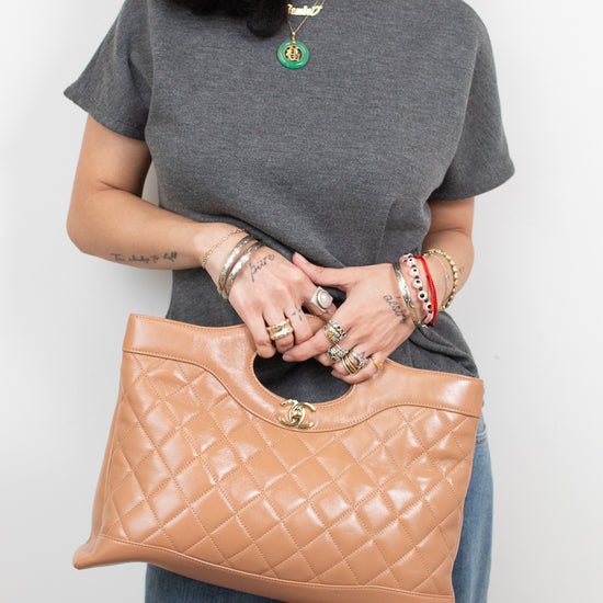 A woman wearing a gray t-shirt and blue jeans holds a tan quilted leather handbag with a gold-tone turn-lock clasp. She is wearing multiple gold rings and bracelets.