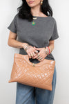 A woman wearing a gray t-shirt and blue jeans holds a tan quilted leather handbag with a gold-tone turn-lock clasp. She is wearing multiple gold rings and bracelets.
