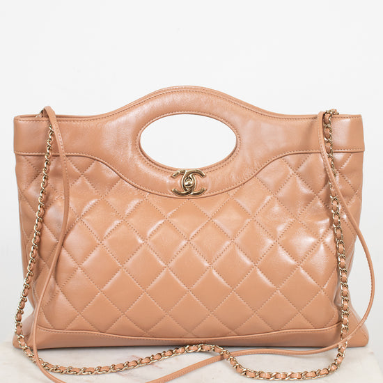 A tan quilted leather handbag with a top handle, gold-tone chain strap, and a small interlocking CC logo on the front, displayed on a light marble surface against a white background.