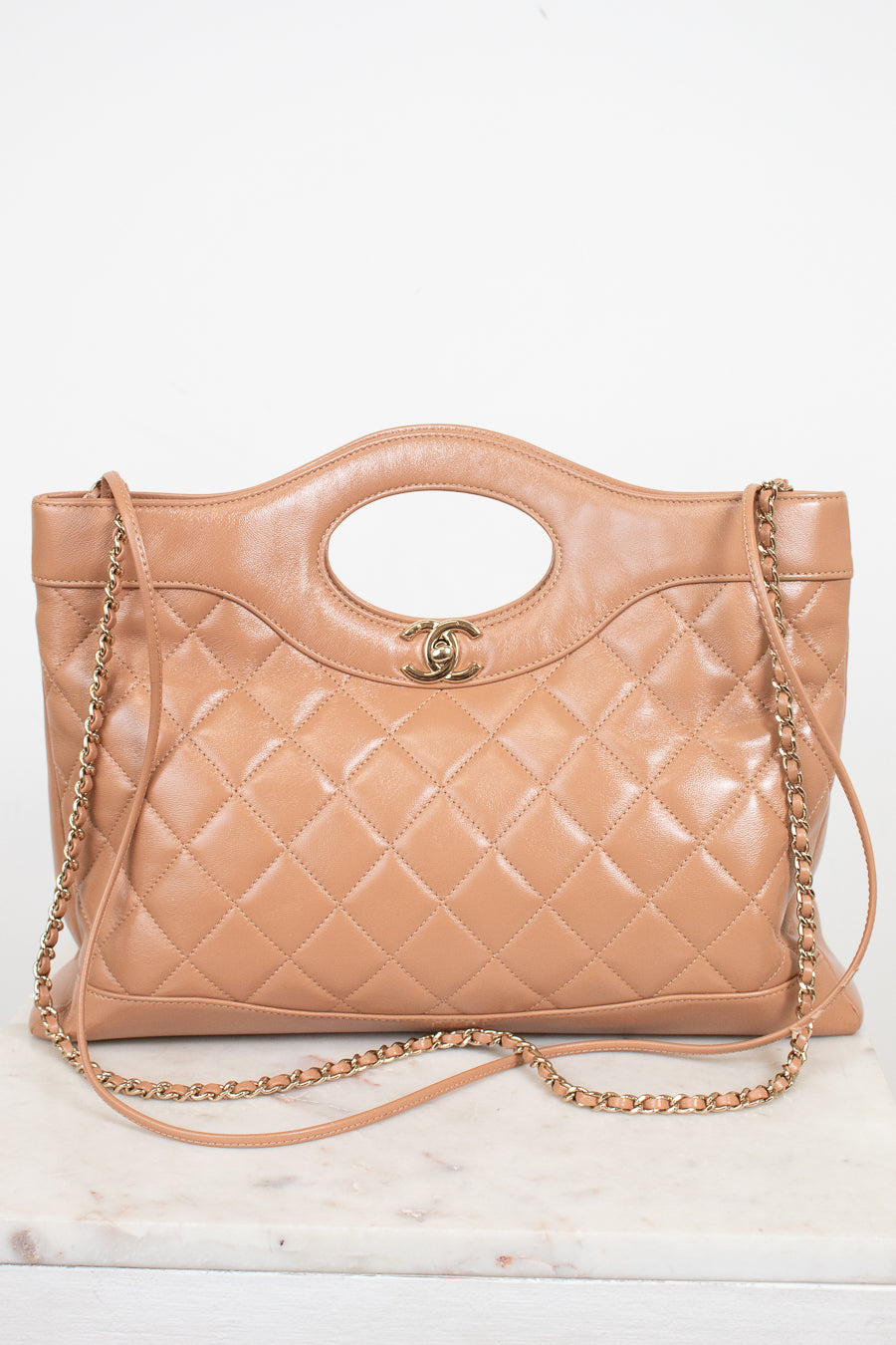 A tan quilted leather handbag with a top handle, gold-tone chain strap, and a small interlocking CC logo on the front, displayed on a light marble surface against a white background.