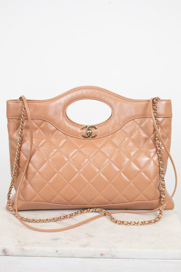A tan quilted leather handbag with a top handle, gold-tone chain strap, and a small interlocking CC logo on the front, displayed on a light marble surface against a white background.