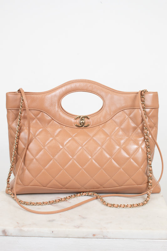 A tan quilted leather handbag with a top handle, gold-tone chain strap, and a small interlocking CC logo on the front, displayed on a light marble surface against a white background.
