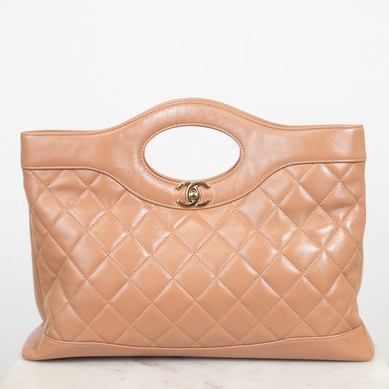 A tan quilted leather handbag with a top handle and a gold interlocking CC logo, displayed on a light marble surface against a white background.