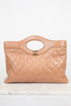 A tan quilted leather handbag with a top handle and a gold interlocking CC logo, displayed on a light marble surface against a white background.