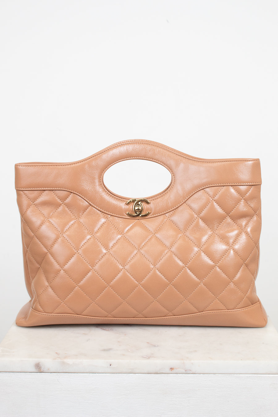 A tan quilted leather handbag with a top handle and a gold interlocking CC logo, displayed on a light marble surface against a white background.