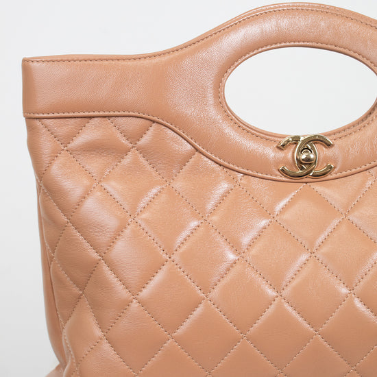 A close-up of a Chanel quilted leather handbag in a warm tan color, featuring a diamond-quilted pattern, a top handle cutout, and a gold-tone interlocking CC logo clasp.