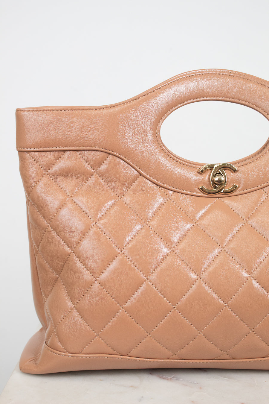 A close-up of a Chanel quilted leather handbag in a warm tan color, featuring a diamond-quilted pattern, a top handle cutout, and a gold-tone interlocking CC logo clasp.