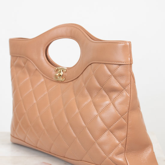 A tan quilted leather handbag with a circular cutout handle and a gold-tone interlocking CC logo, resting on a light marble surface against a white background.